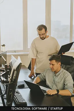 Dois profissionais trabalhando em um ambiente de escritório moderno com múltiplos monitores e notebooks.