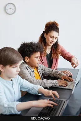 Professora orientando duas crianças que utilizam notebooks em uma sala de aula moderna.