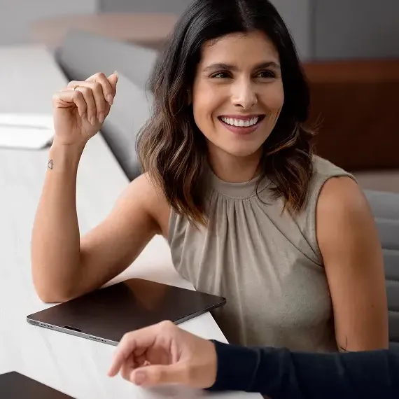Mulher sorridente da equipe LCM Sistemas em reunião, conversando com um colega em uma mesa de escritório branca com tablet.